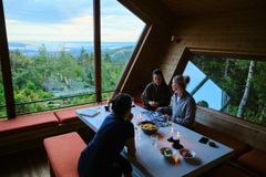 Tre personer sitter ved et bord inne i en moderne hytte, med store vinduer som viser utsikt over naturen utenfor.