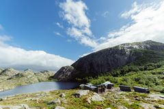 Fjellandskap med en innsjø, hytter og grønne trær under en delvis skyet himmel.