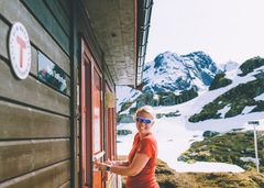 Kvinne som åpner døren til Trollfjorhytta i fjellet, med snødekte topper i bakgrunnen.