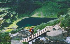 Person som går over en liten bro ved en fjellrygg og grønne omgivelser i bakgrunnen.