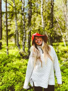 En person i skogen som smiler, iført en rød hatt og hvit genser.