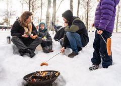 En familie koser seg ved et bål ute i snøen og griller pølser.