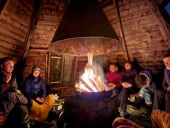 En gruppe mennesker sitter rundt et bål inne i en hytte med trevegger.