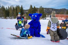 En blå maskot sitter blant barn som leker i snøen på en skibakke.