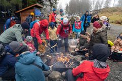 En gruppe mennesker i vinterklær samles rundt et bål for å grille pølser utendørs.