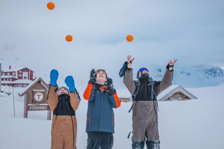 Tre barn leker og kaster applesiner i lufta foran Fondsbu i et snødekket landskap.