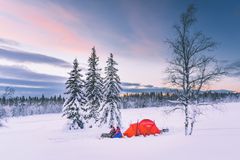 En rød telt i et snødekket vinterlandskap ved siden av snødekte trær, med to personer som sitter foran teltet.