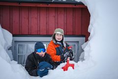 To barn i vinterklær graver i snøen foran en rød hytte med vindu.