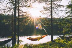 Mennesker som slapper av i hengekøyer blant trær i solnedgang ved en innsjø.