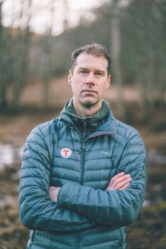 En mann står utendørs med armene i kors, iført en blå dunjakke. Bakgrunnen er uskarp, med skogsområde.