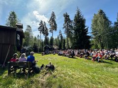 Publikum sitter på benker foran trebygninger, omgitt av skog, under åpningen av en historisk vandrerute på Hedmarken.