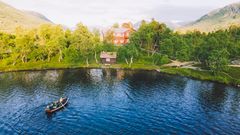 Bilde av en hytte ved en innsjø omgitt av trær og fjell, med en robåt på vannet.