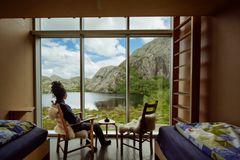 En person sitter i en stol og ser ut av vinduet mot fjell og en innsjø.