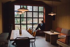 En person sitter og leser ved et stort vindu i en koselig stue med trebord og sofaer.