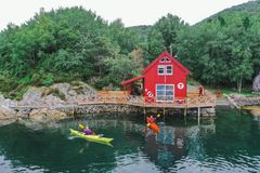 En rød hytte med DNT-logoen ved vannkanten, omringet av trær, med folk i kajakk foran.