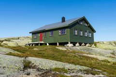 Grønn hytte med røde dører og vinduskarmer, omgitt av steiner og lyng, under en blå himmel.