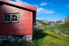 En rød hytte i et fjellandskap i Grasdalen, Ryfylke, omringet av grønn vegetasjon og steiner under en blå himmel.