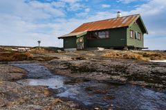 Ei grønn hytte med rustikt tak, omgitt av steinete landskap og blå himmel.