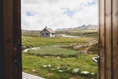 Bilde av en hytte i et fjellandskap med gress, steiner og fjell i bakgrunnen. Hytta er sett gjennom en åpen dør.