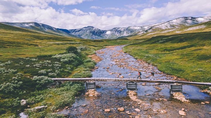 Beskrivelse av bilde: En trebro krysser en steinete elv i et grønt dalføre omgitt av fjell under blå himmel.