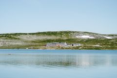 En hytte ligger ved en innsjø omgitt av fjellandskap under en klar blå himmel.