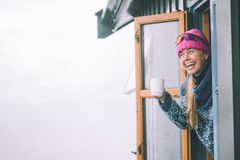 Person med rosa lue og blå genser smiler og holder en kopp mens de ser ut av et vindu