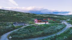 Bilde av Kalhovd turisthytte omgitt av frodig natur og grusveier, med fjell i bakgrunnen.