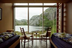 To senger og stoler i en moderne hytte med store vinduer som gir utsikt til fjell og en innsjø.