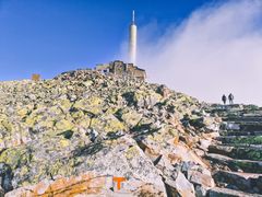 Bilde av Gaustatoppen Turisthytte, en steinbygning nær toppen av Gaustatoppen med steintrapper foran. To personer går på trappene.