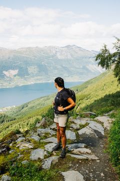 En person med ryggsekk står på en steinete sti og ser utover en innsjø og skogkledde fjell.