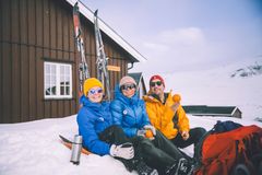 Tre personer sitter i snøen ved en DNT-hytte i fjellet, iført fargerike vinterjakker. De smiler og holder appelsiner. Solrik dag.