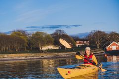 person i en gul kajakk som padler. i bakgrunnen ser man noen hus og skog.