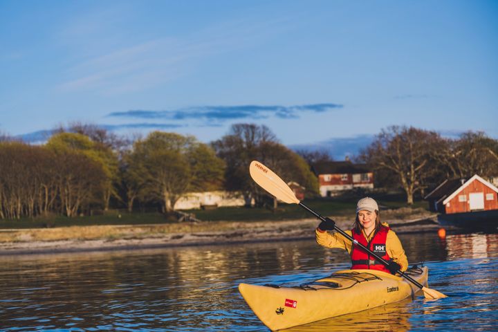 person i en gul kajakk som padler. i bakgrunnen ser man noen hus og skog.