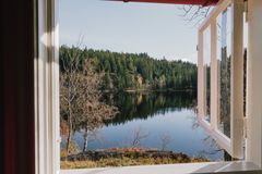 bilde er tatt gjennom et lite vindu i en hytte, og du ser ut over et lite tjern omkranset av grantrær