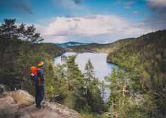 mann med sekk og regnbuelue som står på en høyde og ser utover et vann og skog