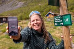 Kvinne smiler og holder frem en smarttelefon ved siden av et SjekkUT-skilt i fjellandskap.
