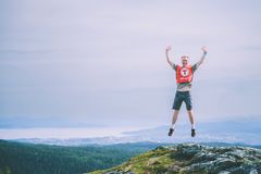 En person hopper på en fjelltopp iført en rød ryggsekk. Utsikt over en by og vann i bakgrunnen.