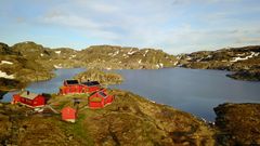 En samling av røde hytter ved en innsjø i et fjellandskap med snøflekker i Ryfylke.