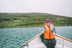Kvinne med oransje redningsvest ror en båt på en innsjø, med fjelltopp og hytter i bakgrunnen.
