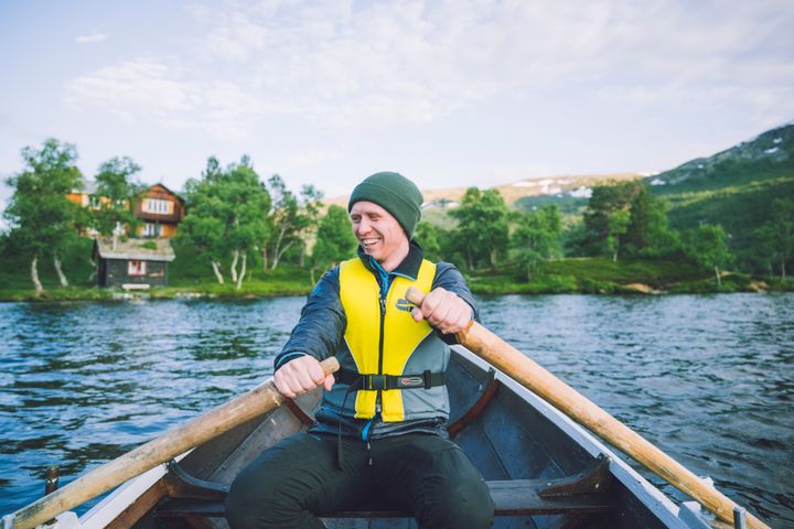 En person i redningsvest ror i en robåt på en innsjø, med natur og en hytte i bakgrunnen.
