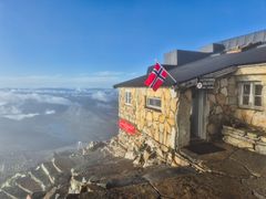Steinbygning ved Gaustatoppen med et norsk flagg, omringet av skyer og fjellandskap.