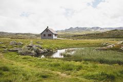 En enslig hytte i et grønt landskap med steiner og en liten dam i nærheten, omgitt av fjell i det fjerne.
