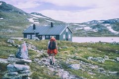 En person med ryggsekk går på en sti mot en hytte i fjellet, med snødekte fjell i bakgrunnen.