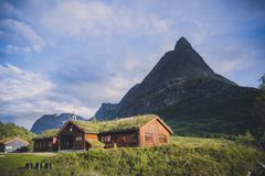 Rustikk hytte med gressdekket tak i frodig landskap, fjell i bakgrunnen.