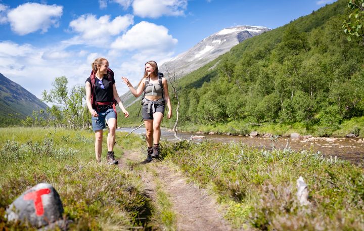 To personer går tur i fjellet langs en sti i solfylt vær, med en rød T-merket stein i forgrunnen.