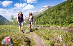 To kvinner vandrer langs en sti i Trollheimen en solrik dag, med fjell i bakgrunnen. En stein ved stien er merket med en rød T.
