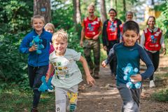 En gruppe glade barn løper på en skogsti, noen holder små blå kosedyr. Voksne går bak dem i røde vester.