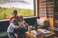 Mor med to barn sitter på en sofa i en koselig hytte med utsikt over naturen gjennom et stort vindu.