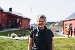 Smilende kvinne står utendørs mellom røde bygninger ved vannet.