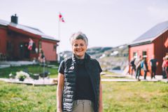 En smilende kvinne med kort grått hår står ute på en gressplen, med røde trehus og et norsk flagg i bakgrunnen.